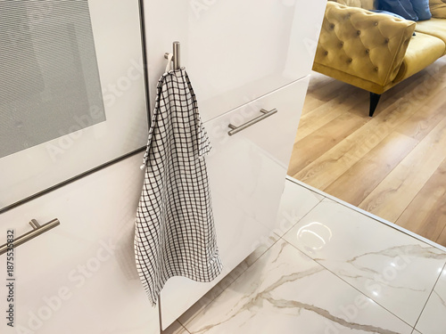 A checkered kitchen towel is hanging on a metal handle of a white kitchen cabinet, with a kitchen floor and furniture visible in a modern interior.
