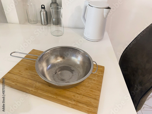 A stainless steel metal colander is placed on a wooden cutting board on a kitchen countertop, with kitchen utensils in the background.