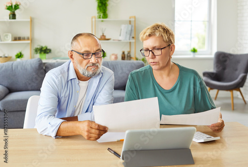 Senior couple sits at table with tablet and paperwork, discussing budget, expenses and retirement plans checking online accounts. Elderly man and woman review bills and manage finances together