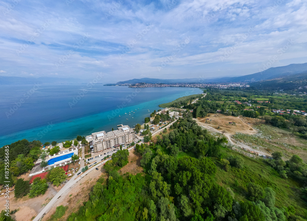 Obraz premium Aerial drone view of the scenic Lake Ohrid waterfront in North Macedonia, featuring red-roofed buildings, lakeside hotels, and the clear blue waters of this UNESCO World Heritage site.