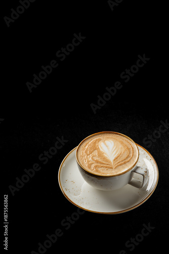A white cup of hot cappuccino with beautiful heart-shaped latte art is presented on a matching saucer against a black background, with copy space available
