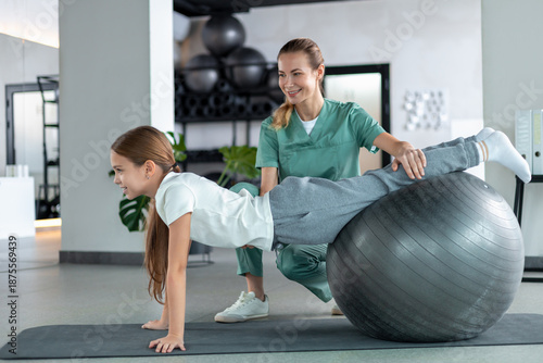 Картината върху платно Little girl holding plank on fitness ball with female therapist assisting