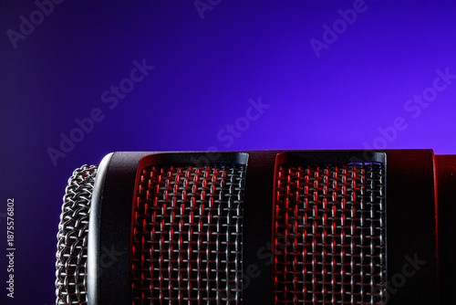 A microphone sits on a table with a dark background. The mesh part reflects red and purple light. The scene shows a recording session setup. Equipment is ready for use.