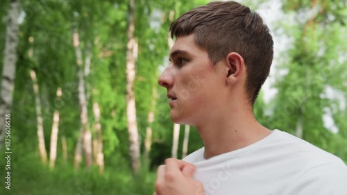 Side view of big brother standing in summer forest holding white polo collar and fanning himself, showing heat discomfort, warmth, and natural light expression in green outdoor environment