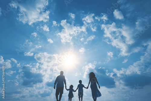 A family of four walks together holding hands against a bright sun and blue sky with clouds