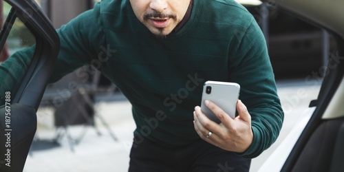 Man with smartphone leaning into car, wearing green sweater. Focused on phone, the man appears engaged. Car, smartphone, and man are central elements. A man using a smartphone to get a car.