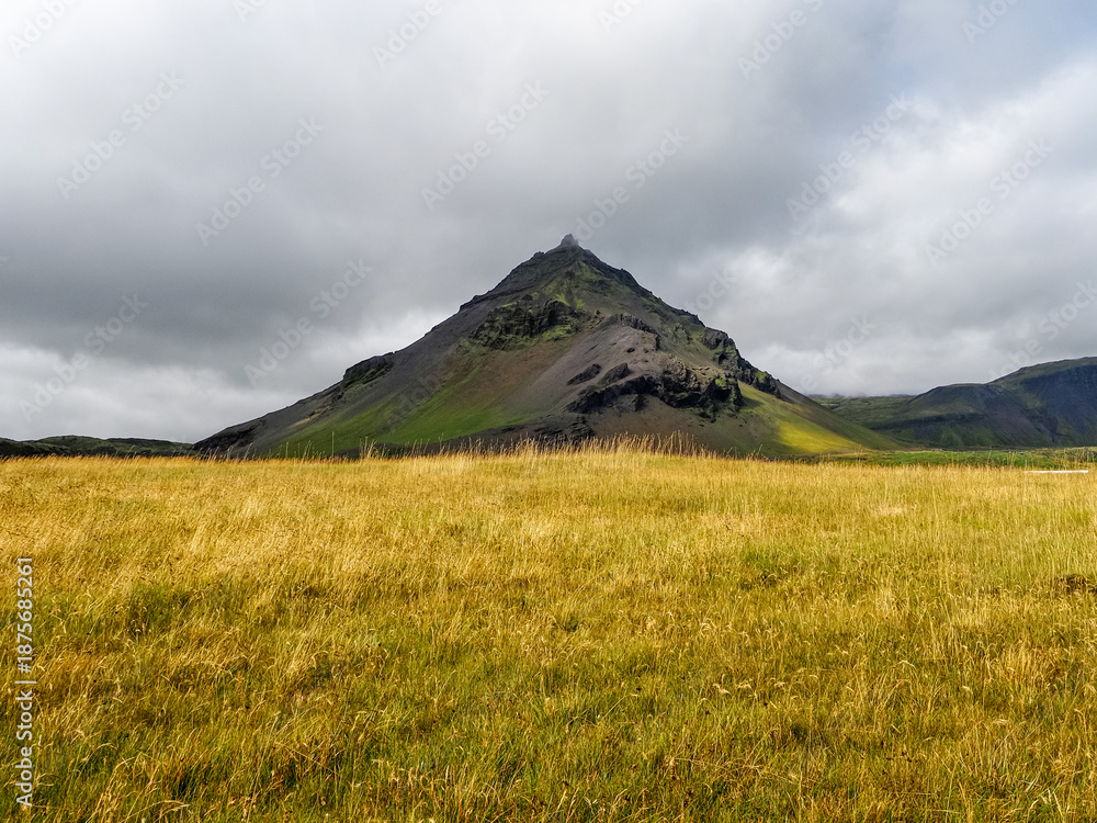 Fototapeta premium Wild Icelandic landscape with visible Stapafell volcanic mountain, against dark cloudy sky. This palagonitic pyramide is 526 m in height. Hellnar, Arnarstapi, Snæfellsnes Peninsula in western Iceland.