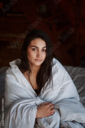 A woman with black hair wakes up in the morning in a white blanket and catches the rays of the sun. The portrait is creative, with brown eyes. High quality photo