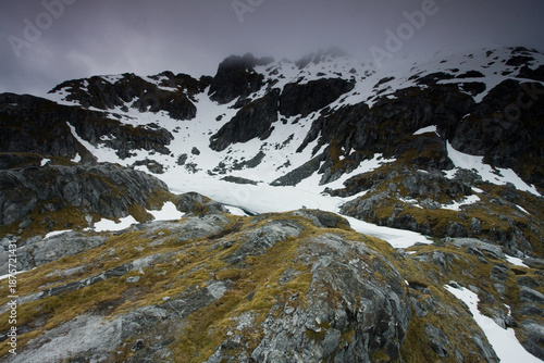 View of rugged terrain where moss-covered rocks meet snowy slopes beneath a brooding sky, a stark contrast of textures and tones, Christchurch, West Coast Region, New Zealand.