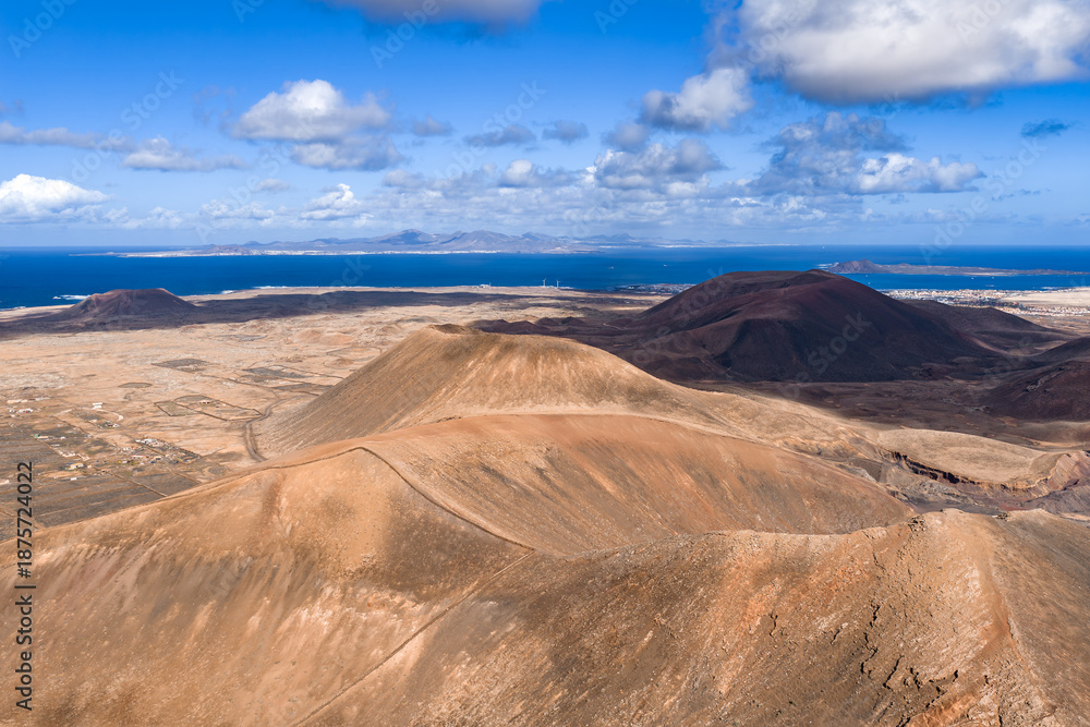 Naklejka premium Aerial scene shows Fuerteventura cones and calderas near Caldera Los Arrabales and Playa del Bajo de la Burra, Lanzarote outline, coastal town, and salt white beaches under midday light.