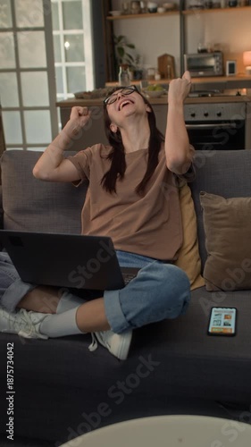 Vertical shot of young biracial girl resting at home on sofa while making online payment on laptop using credit card and waving euphorically celebrating great sale