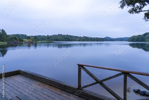 View of the tranquil lake mirroring the overcast sky, observed from a weathered wooden dock under the shade of trees, Treungen, Telemark, Norway.
