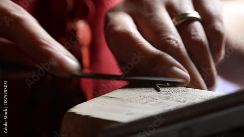 Close-up view of artisan hands filing a metal jewelry piece on a wooden bench pin. The craftsperson carefully shapes the object using a hand file in a bright workshop.

