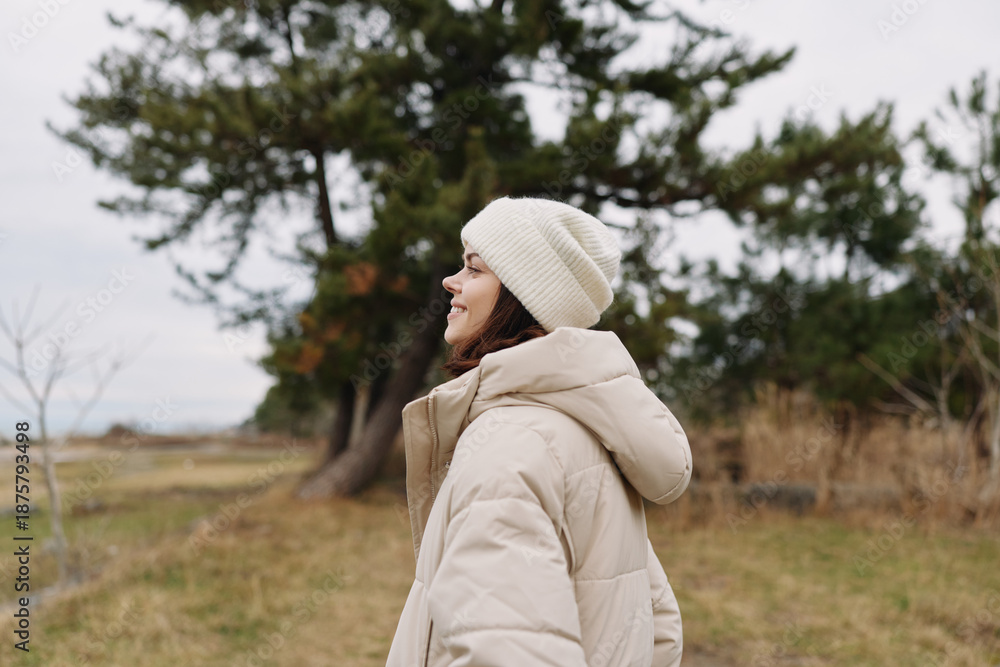 Fototapeta premium Outdoor scene with a person wearing a beige winter coat and knit beanie, standing in a park among trees and open field, calm weather and reflective mood.