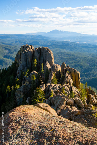 A shaded dirt path winds through a lush forest of aspen and ponderosa pine on the Devil's Head Lookout Trail in Colorado, with massive granite boulders and glimpses of the rugged Rampart Range peaks.