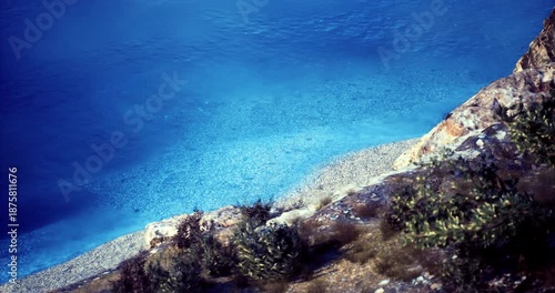 Clear turquoise waters gently lap against a pebbled beach surrounded by lush greenery. Sunlight dances on the water, creating a serene atmosphere perfect for relaxation.