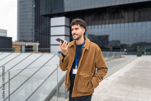 Young man standing outdoors in an urban setting during a work break, dictating a voice message on his smartphone while smiling