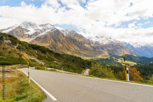 An asphalt road on the San Bernardino Pass in Switzerland The mountains are partially covered in snow. The mountains are also covered in a beautiful, colorful autumn blanket sky is blue and cloudy