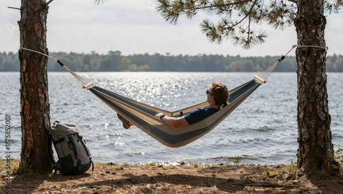 A person relaxing in a hammock strung between two pine trees at a lakeside campsite, with a backpack resting nearby