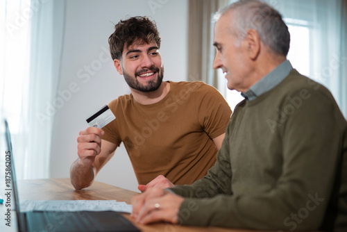 A young man assisting his senior father with online banking and card payment at home. Two generations discussing finances, bills, and digital payments in a comfortable home environment. Family support