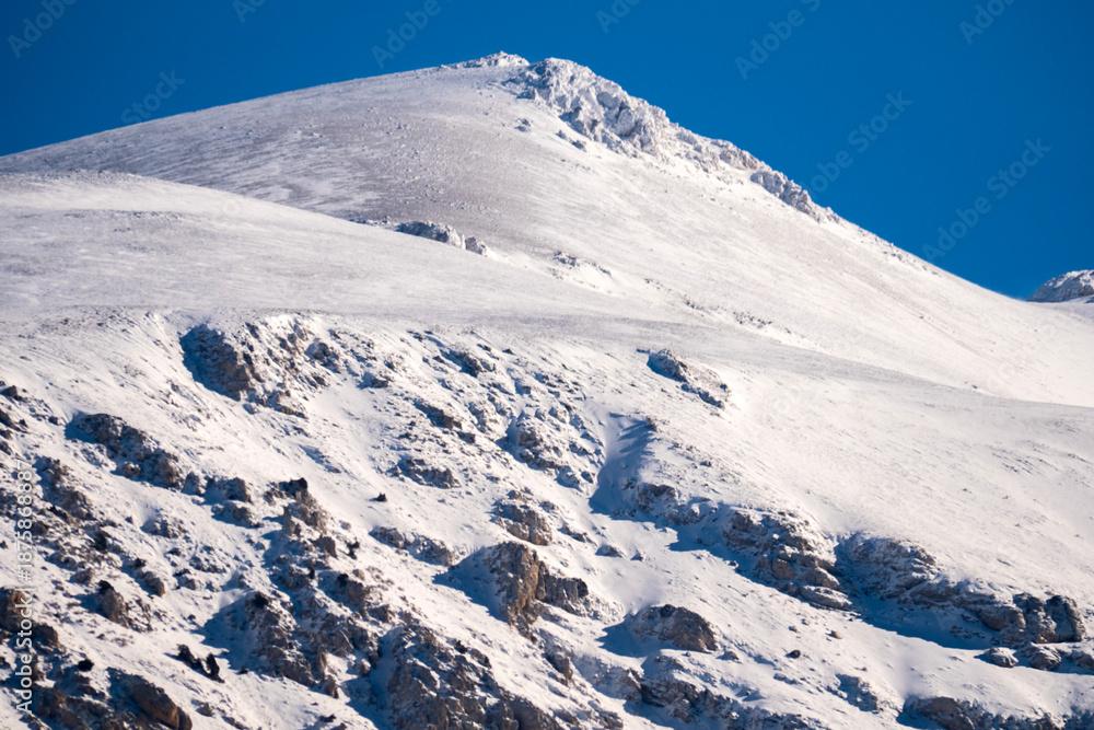 Obraz premium Majestic snowy mountain summit reaches into the clear blue sky under the bright winter sun featuring dramatic shadows and pristine white snow textures in a wild nature scene