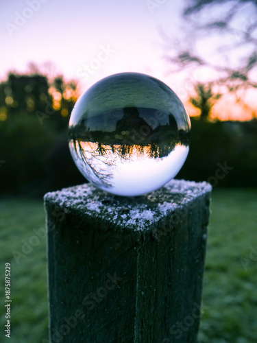 A glass ball is sitting on a wooden post in a field. The ball is reflecting the sky and the surrounding trees in a mirror-like effect. The scene is serene and peaceful
