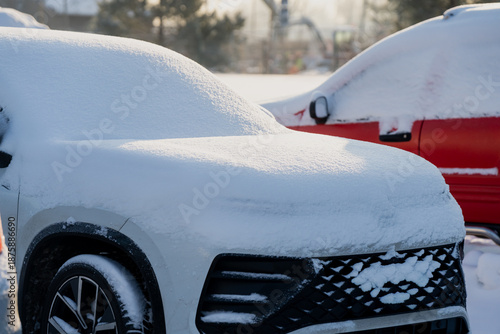 Parked SUV completely covered in fresh snow, front grille and wheel partly visible, winter parking lot scene with red snow covered car blurred in background