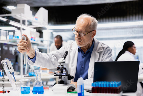 Senior lab manager pipetting agent into blue test tube sample to improve chemical formulation efficiency. Elderly man observing reaction in test tube to verify scientific hypothesis