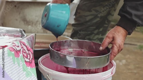 An unrecognizable elderly winemaker pours red grape juice through a sieve into a bucket.