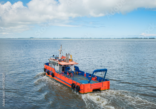 Industrial orange workboat sailing on calm blue sea under a cloudy sky. Rear view of a professional service vessel navigating open coastal waters