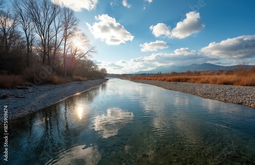 Wide river flows through dry grass banks under a blue sky with fluffy clouds. Bare trees line the shore, sun reflects on water surface. Distant mountains are visible on horizon.