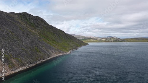 Aerial view of a high rocky slope above a narrow green water edge in Russia, Chukotka, Lavrentiya Bay at daytime under broken cloud. Minimal wake and clear color serve premium travel need