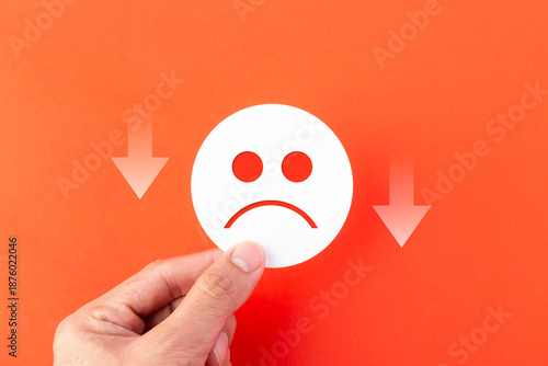 A hand holding a white sad face emoji icon on an orange background with downward arrows. Concept for business failure, customer dissatisfaction, negative feedback, and economic decline.