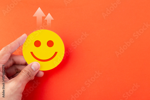 A hand holding a yellow happy face emoji icon on an orange background with upward arrows. Concept for business growth, customer satisfaction, positive feedback, and financial success.