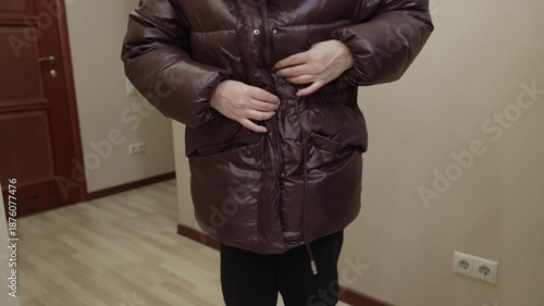 Adult woman standing indoors fastening brown winter jacket with hands, preparing to go outside. Cold season clothing, daily routine and lifestyle concept.