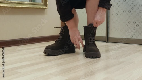Adult woman standing indoors putting dark boots on bare feet on wooden floor. Getting ready, footwear and daily routine concept.