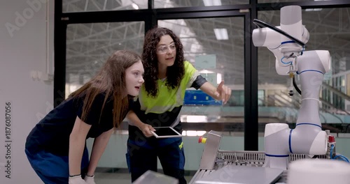 Female trainee reacting with excitement while observing robotic arm operation during slowmotion scene in industrial automation lab for smart factory training under supervision of asian instructor