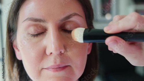 Closeup of a mature woman with closed eyes while a makeup artist applies liquid foundation with a brush in a professional beauty salon. Skincare, cosmetics, wellness, pampering concept