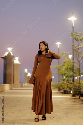 Stylish young woman wearing a brown long maxi dress posing confidently on an illuminated urban walkway at night