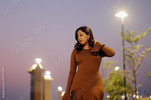 Stylish young woman wearing a brown long maxi dress posing confidently on an illuminated urban walkway at night