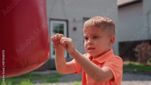 Child Strikes Red Bag Happily, Young Boy Enthusiastically Punches Bright Red Bag In Sunlight, Energetic Caucasian Boy Delivers Playful Punches To Colorful Bag Outdoors In Sunny Yard