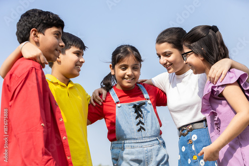 Happy group of indian kids friends embracing and sharing a candid conversation together in summer park, concept of teamwork and friendship. Children having fun talking and discussing outdoor.