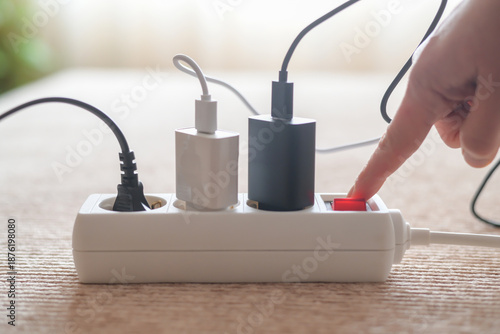 Human hand is switching off a white power strip, with multiple plugs and chargers connected, illustrating the concept of saving energy and reducing electricity consumption