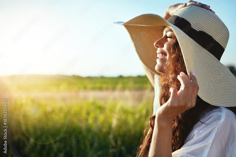 Fototapeta premium Smile, woman and hat for travel in nature for fresh air, enjoy sunshine and summer holiday. Space, female person and meadow for peace, calm morning and vacation trip for stress relief in countryside
