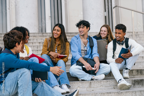 Group of diverse university students smiling and chatting happily while sitting on campus stairs outside the building