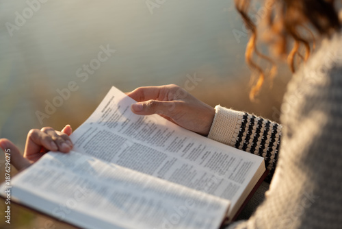 The image depicts a woman reading the Bible outdoors in soft sunlight. The concept encompasses reading the Bible and seeking God's words in the sacred texts, spiritual growth, and daily devotion. © Wanan