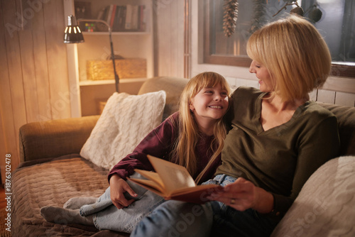 Schilderij op canvas A woman reads a book to a smiling girl while they sit together on a sofa