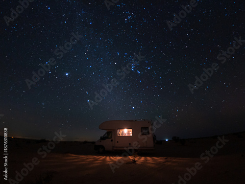 Night scene in the Douz desert, Tunisia, with a camper van illuminated from inside beneath a star-filled sky featuring the Orion constellation and bright Jupiter, creating a serene adventure mood