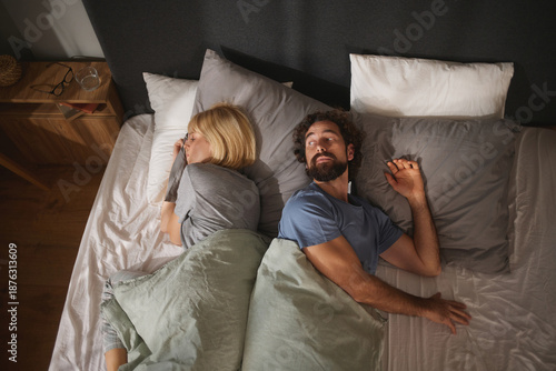 Couple in bed lies back-to-back, showing signs of emotional distance and unresolved relationship problems. The dim lighting enhances their feelings of disconnect and tension.