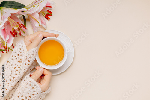 Female hands hold cup of tea with gentle lily flowers and copy space for product presentation or design, beige background, top view